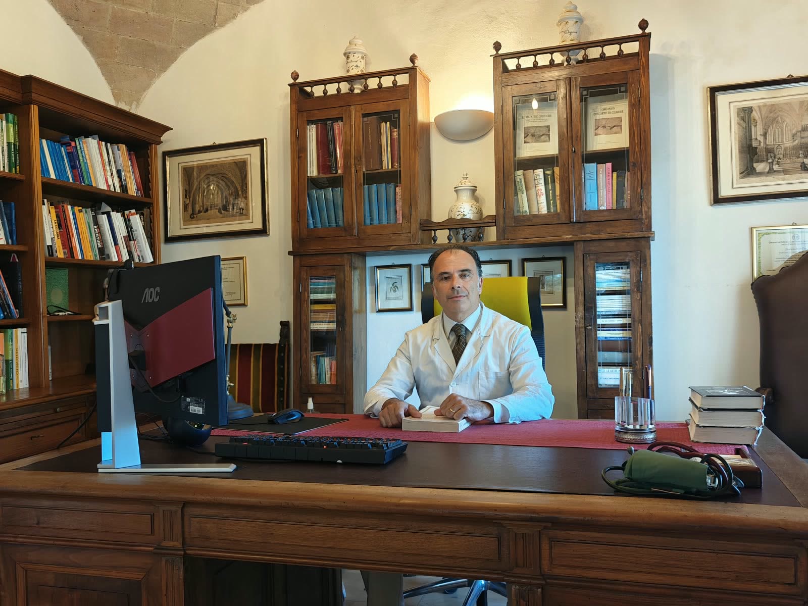 Il Dott. Tancredi Ascani in camice bianco alla scrivania del suo studio, davanti a vetrine in legno con libri e antichi vasi da farmacia.